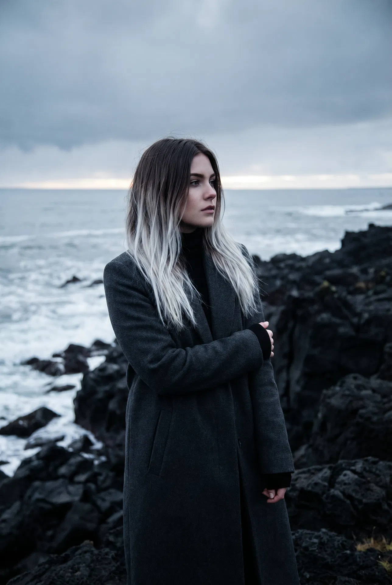 Ikat — Zora Wisp Weft model on Icelandic volcanic coastline, charcoal coat, dark-root-to-platinum melt